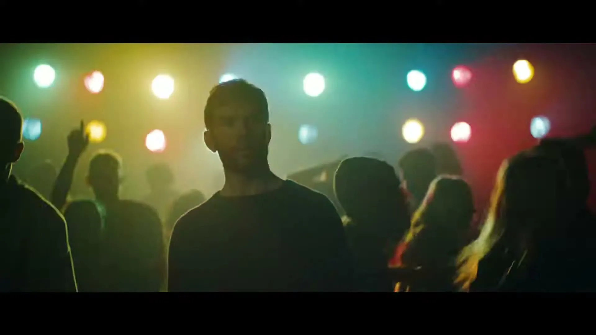 A young man with short hair looks around a crowded, dark club with colorful lights flashing in the background, as people dance around him.