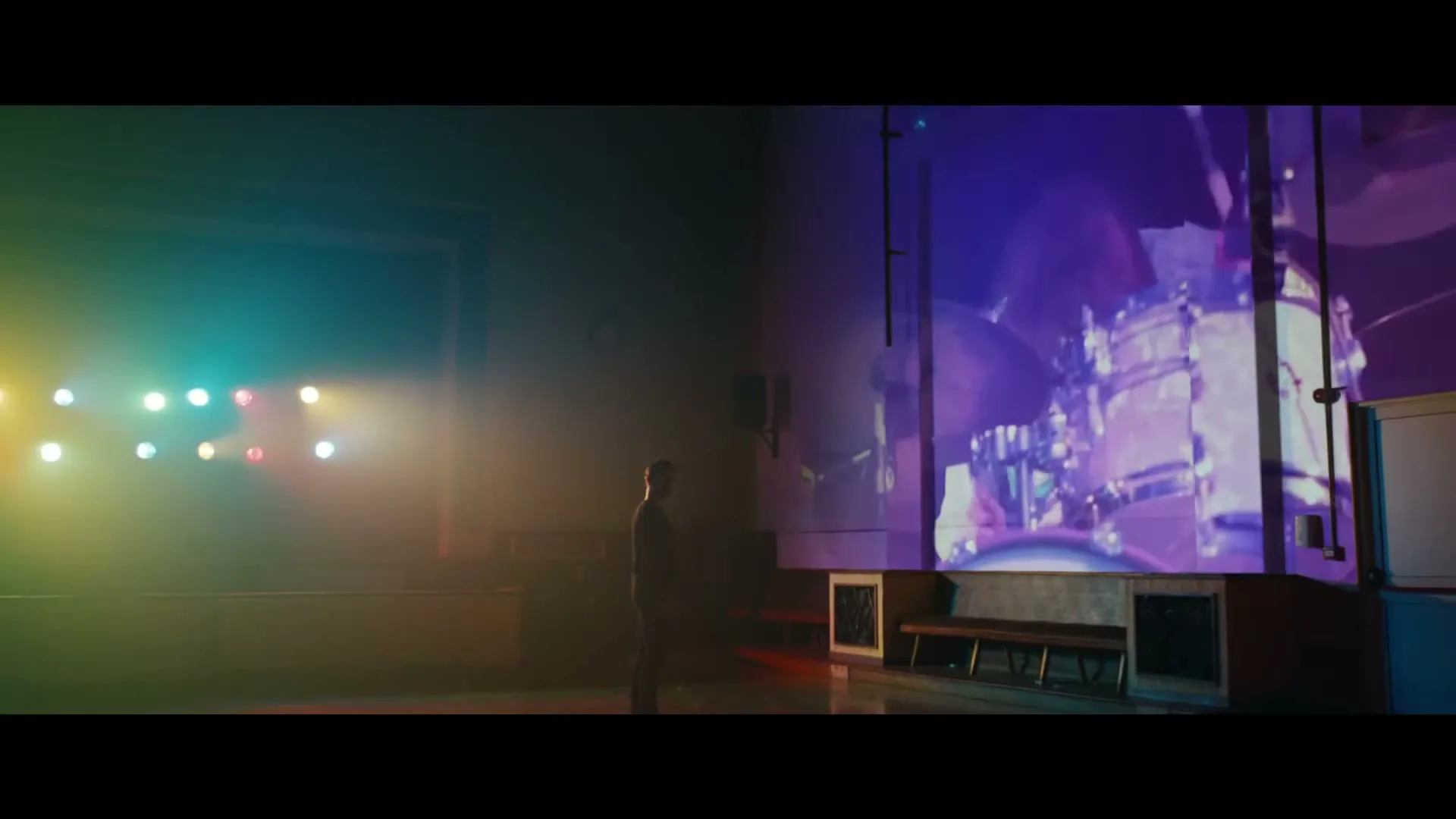 A man stands in a dark hall, watching a large screen displaying a drummer playing, with colorful stage lights illuminating the background.