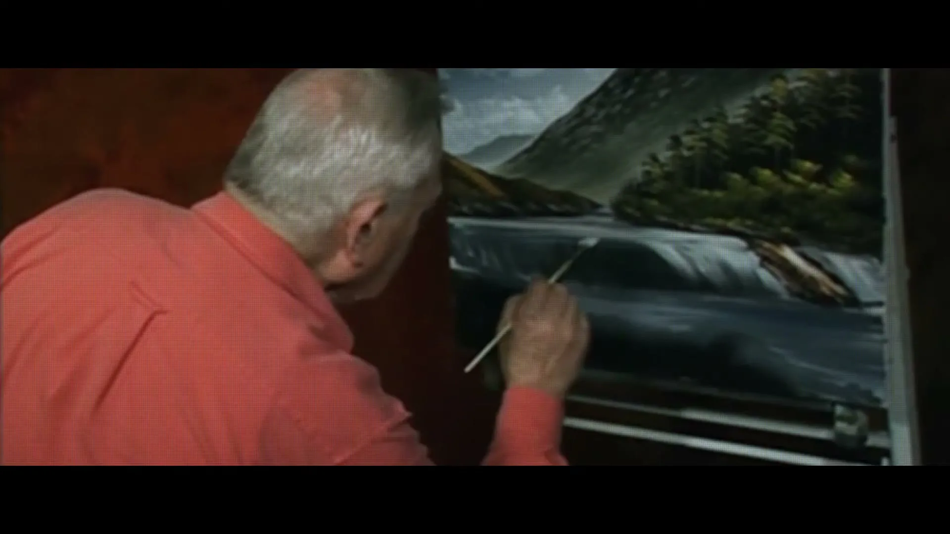 A man with gray hair and a red shirt paints a landscape with a waterfall on a canvas in an interior setting.
