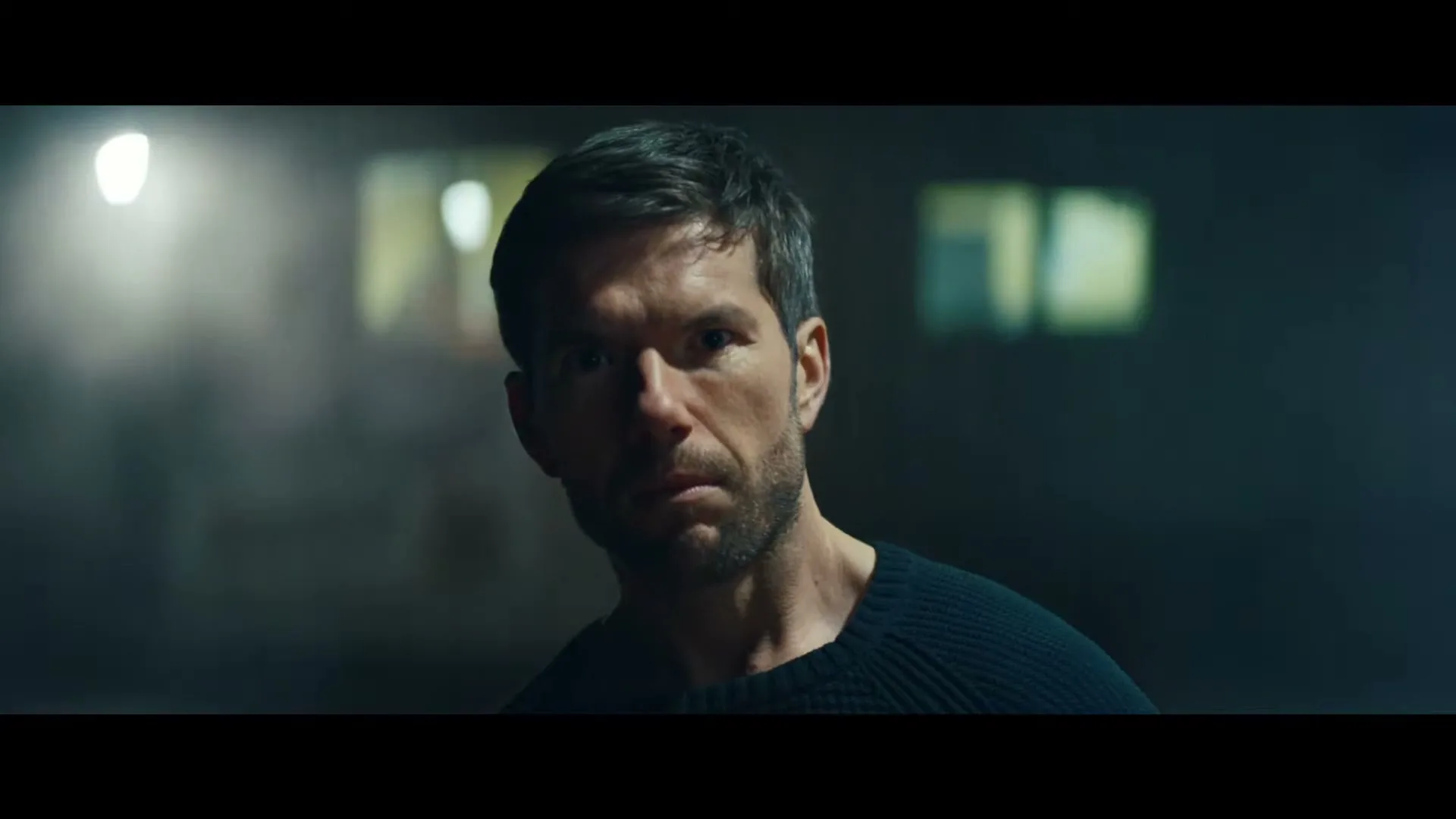 A man with short dark hair and a beard looks intently at the camera in a dimly lit, industrial-looking interior.