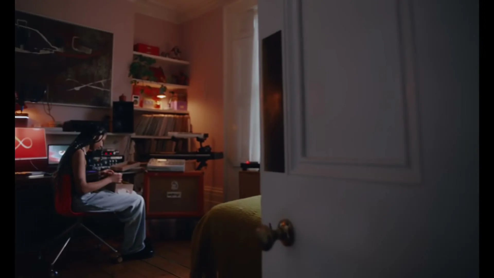 A young woman with braids sits at a desk in a bedroom, focused on a music mixing board and computer screen, as a white door slowly opens to reveal her.
