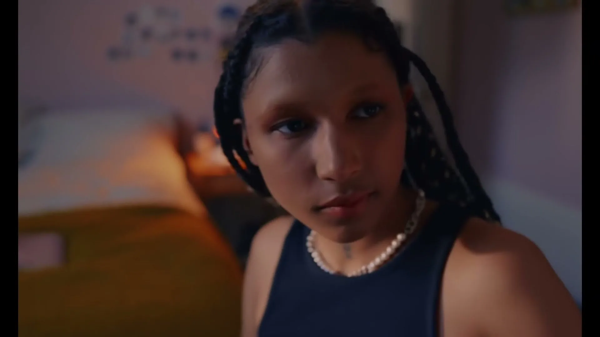 A young woman with braided hair and a pearl necklace looks up from what she is doing, in a dimly lit bedroom.