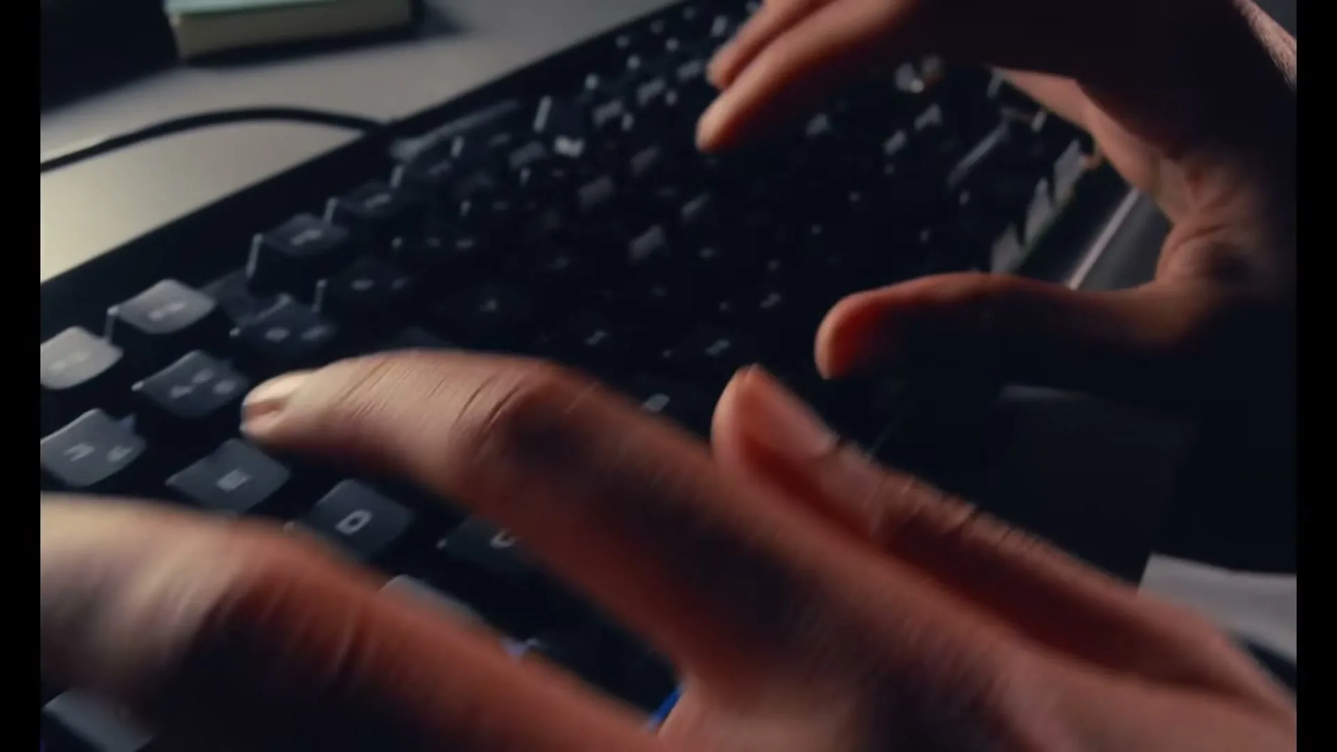 A person's hands rapidly type on a black computer keyboard with blue backlighting in a dimly lit room.