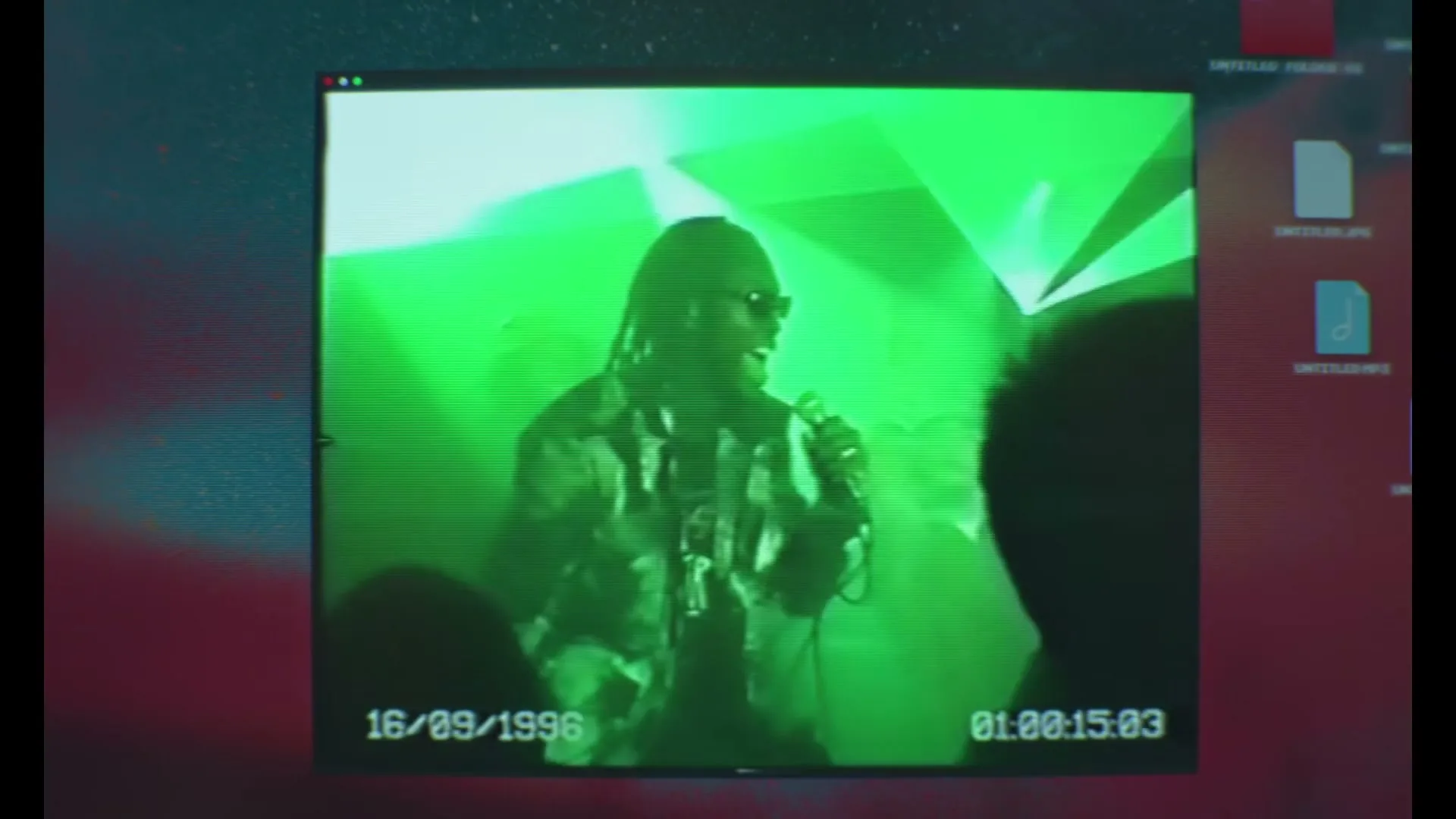 A person with dreadlocks and sunglasses performs on stage under green lights, viewed through a retro computer screen interface with a date and time stamp.