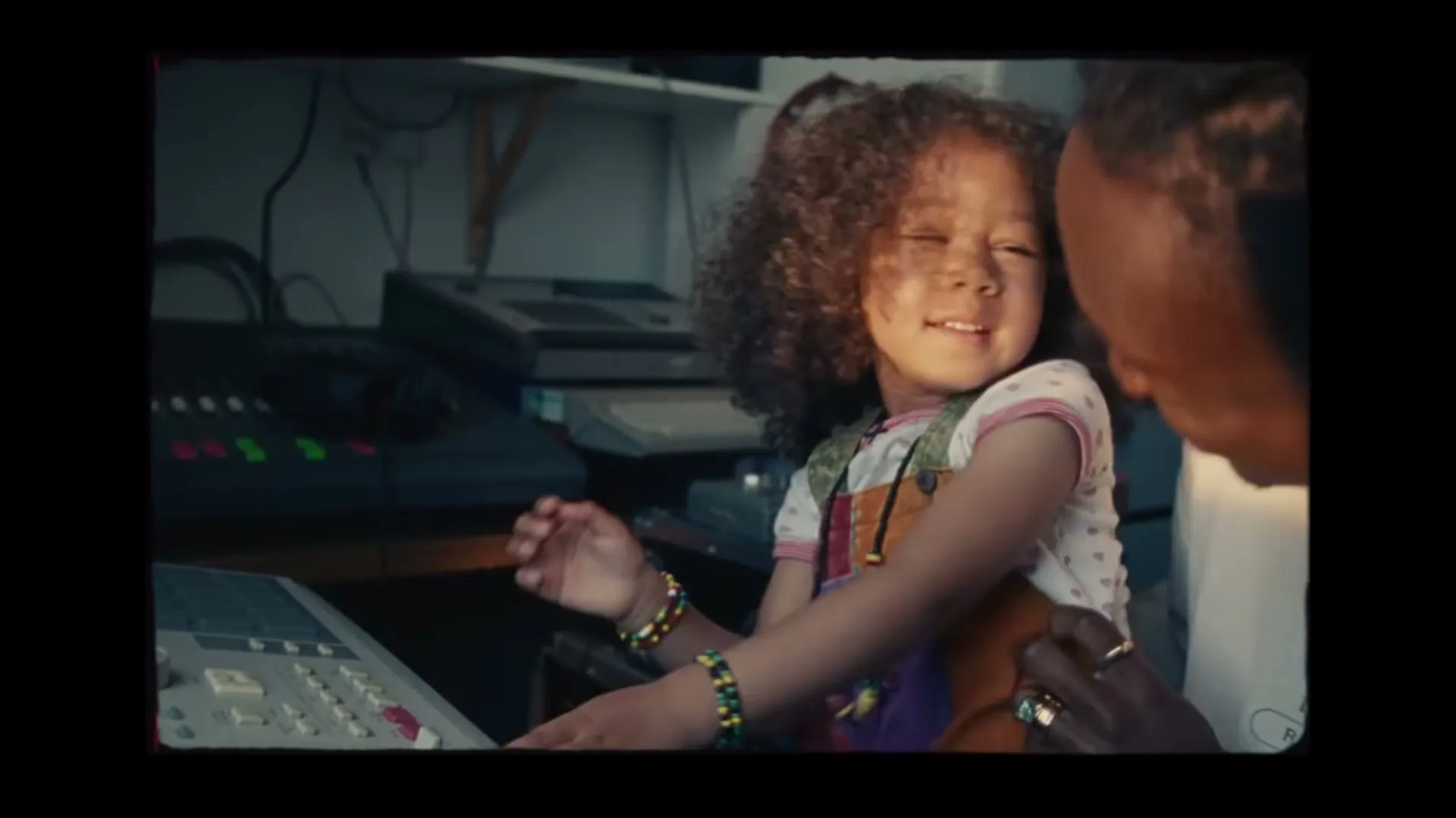 A young girl with curly hair sits on an adult's lap, smiling and playing with a sound mixing board in a home studio.