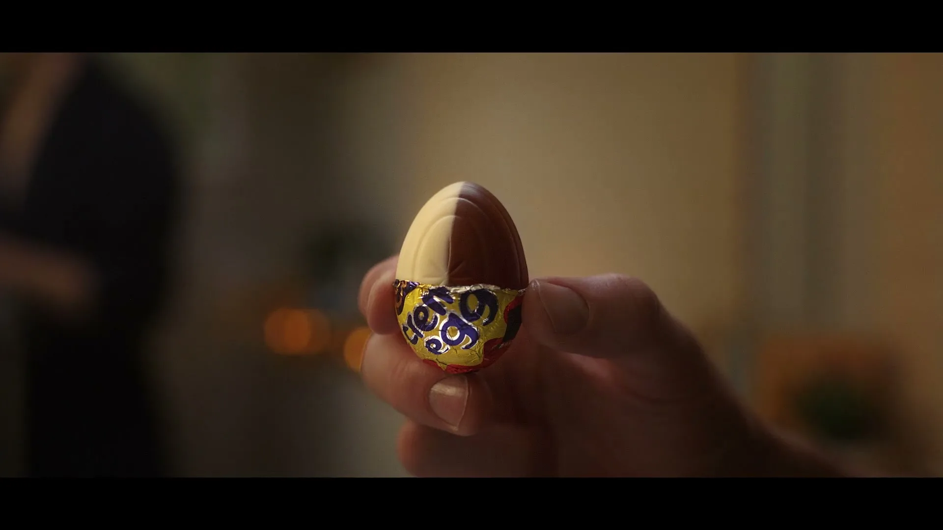 A hand holds a half-unwrapped Cadbury Creme Egg, showing the chocolate and cream filling, in a softly lit interior setting.
