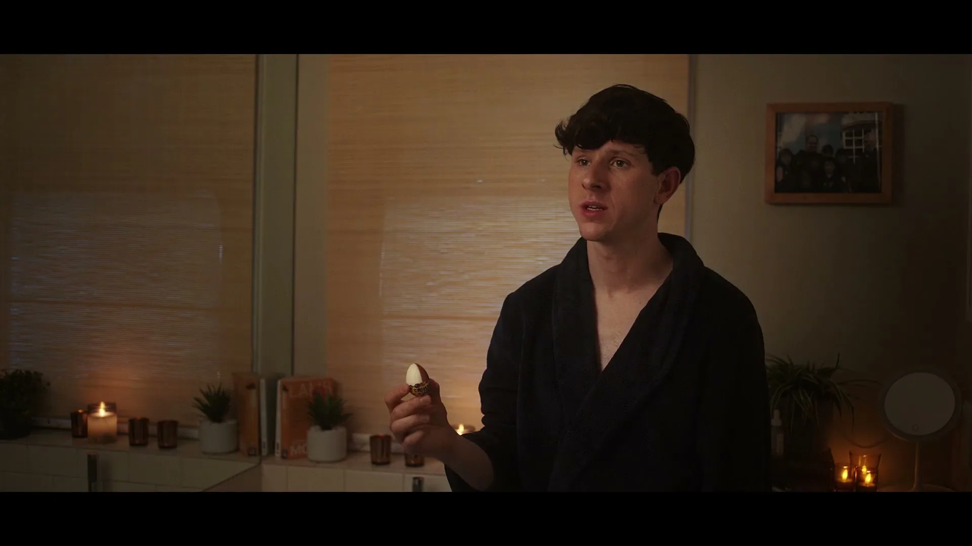 A young man in a dark robe holds a small, golden egg-shaped object, looking at it with a curious expression in a dimly lit room.