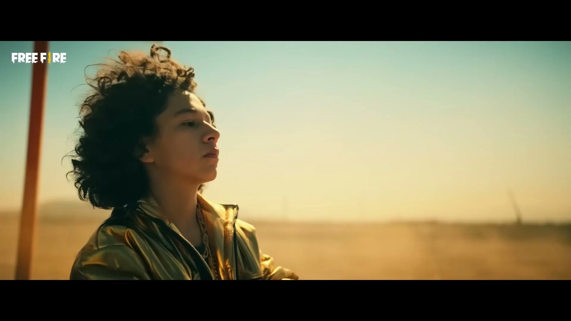 A young person with curly hair and a gold jacket looks off into the distance in a desert landscape under a bright sky.