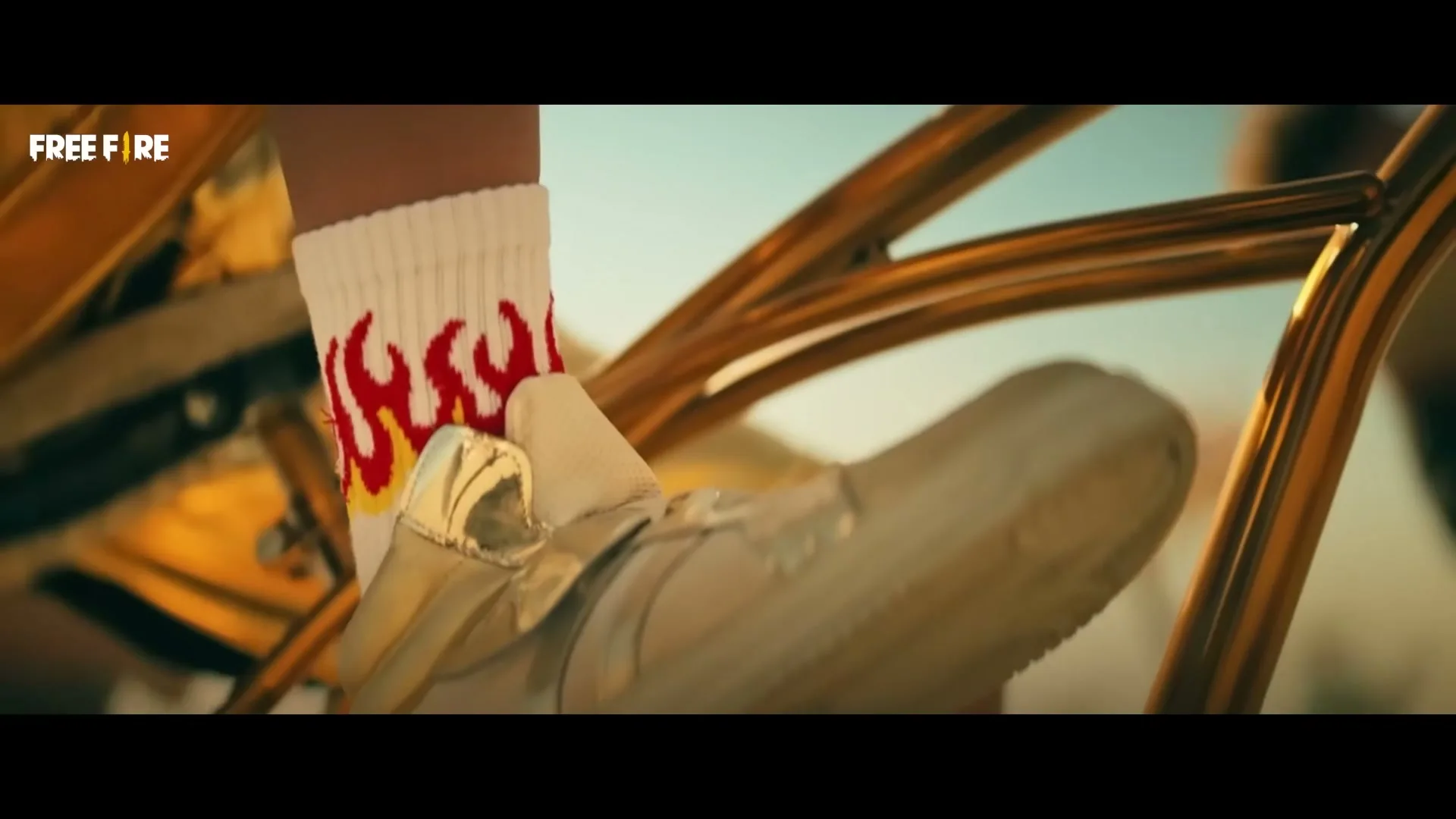 A person's foot in a white sock with red flames and a gold shoe rests on a gold bicycle pedal in an outdoor setting.