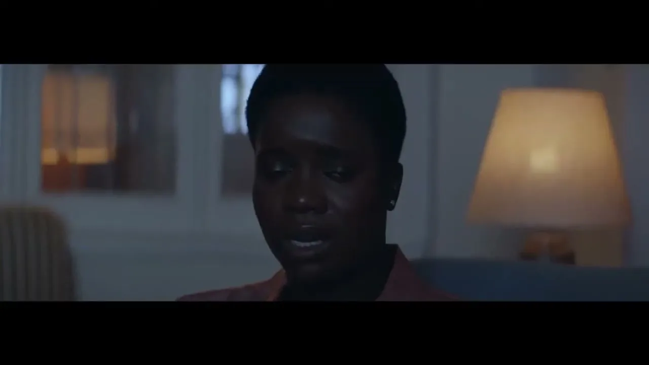 A young Black woman with short hair, wearing a pink top, looks distressed while sitting on a couch in a dimly lit living room at night.