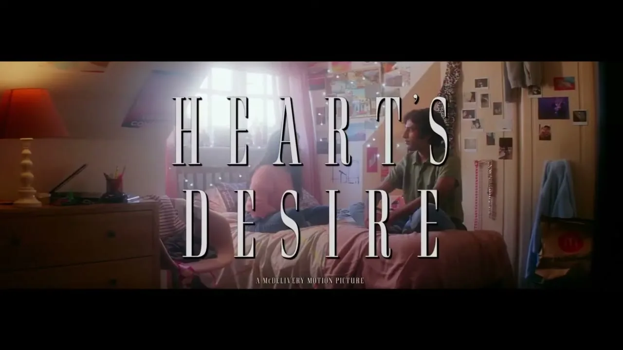 A young couple sits on a bed in a cozy bedroom, looking at each other, with a movie title card superimposed over the scene.