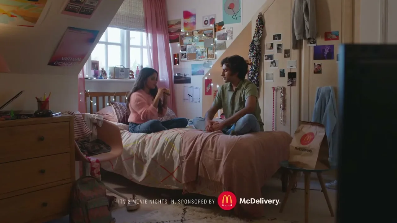 A young woman and man sit on a bed in a cozy, decorated bedroom, eating McDonald's from a paper bag.