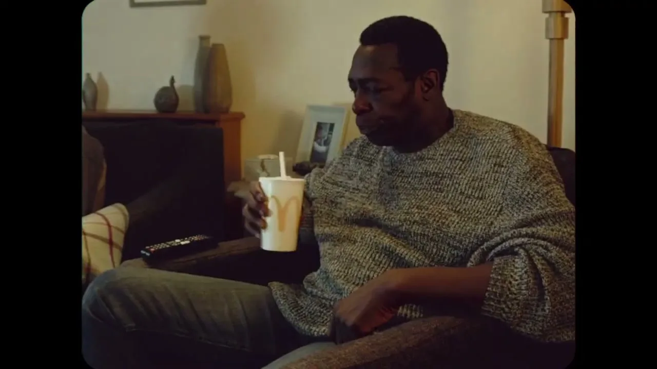 A man in his 30s sits on a couch, drinking from a McDonald's cup in a cozy living room.