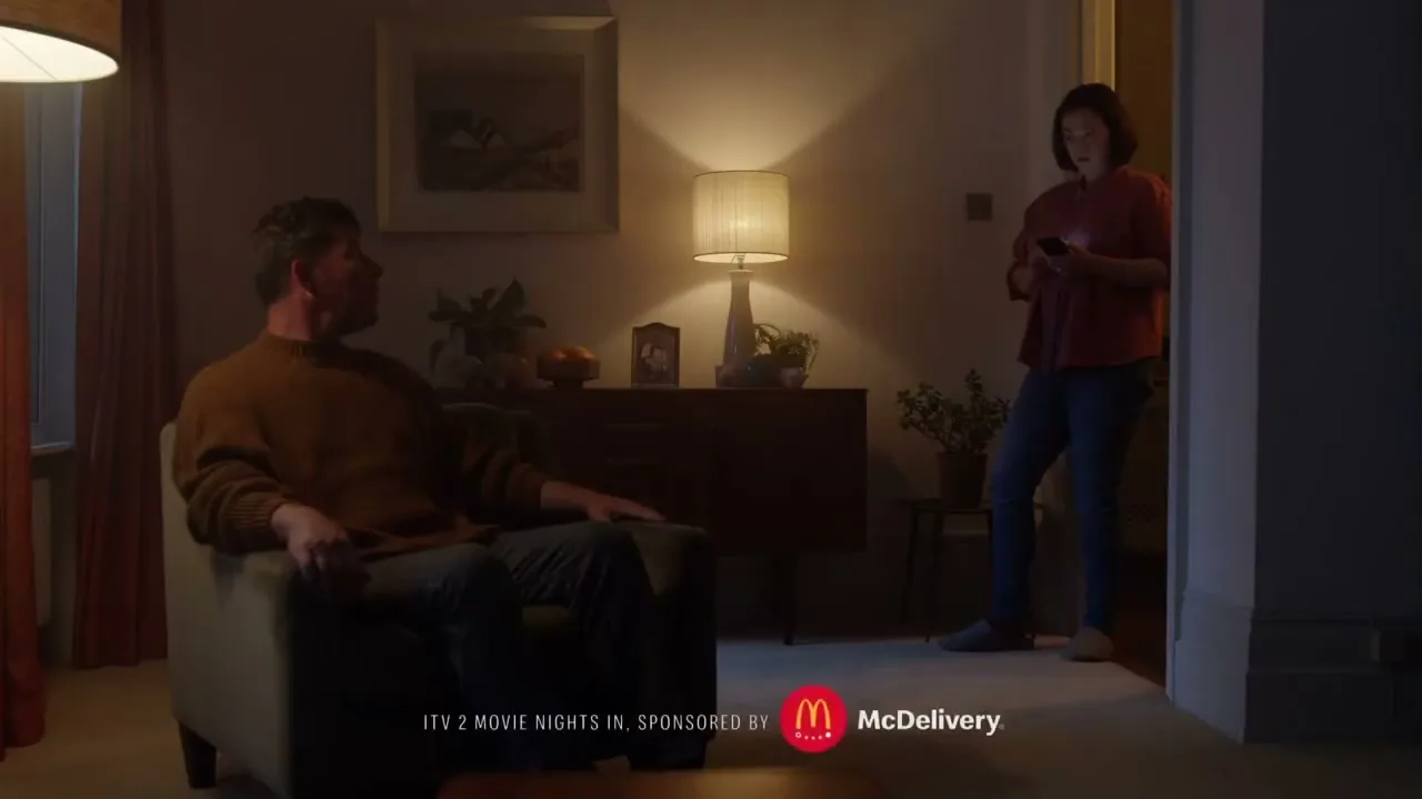 A man sits in an armchair while a woman stands in a doorway, both looking at each other in a dimly lit living room.