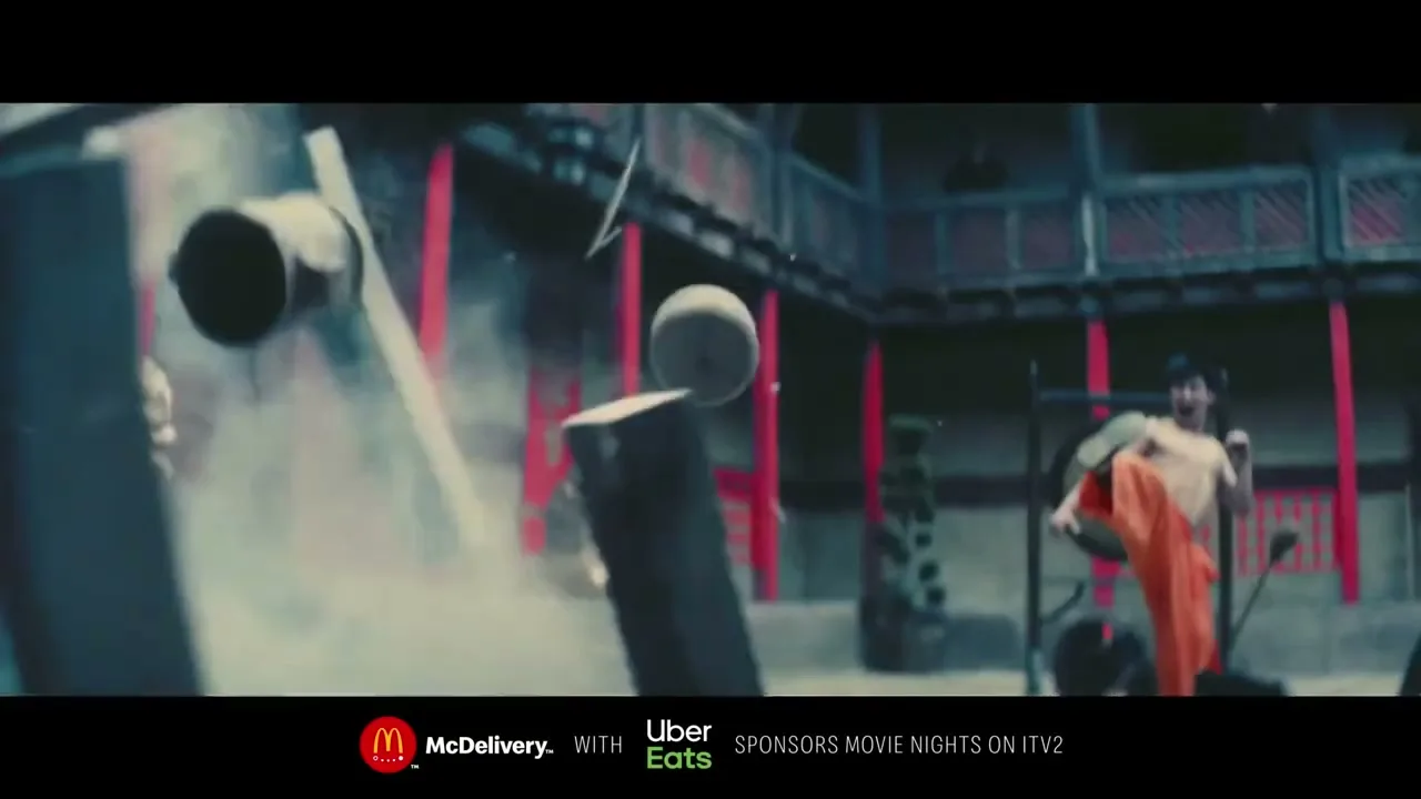 A young man in orange pants practices martial arts, kicking a wooden dummy that breaks apart in a courtyard.