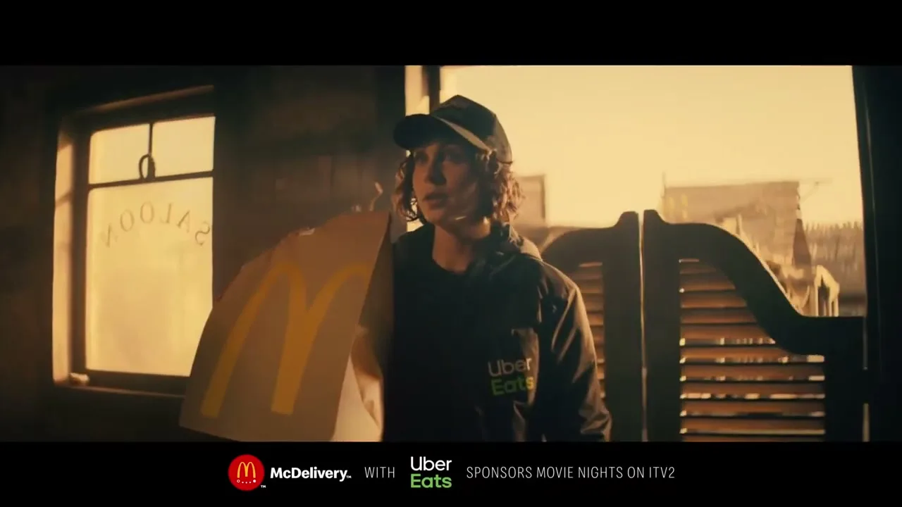 A young delivery person in an Uber Eats uniform stands in a dimly lit saloon, holding a McDonald's bag, with bright light from outside.