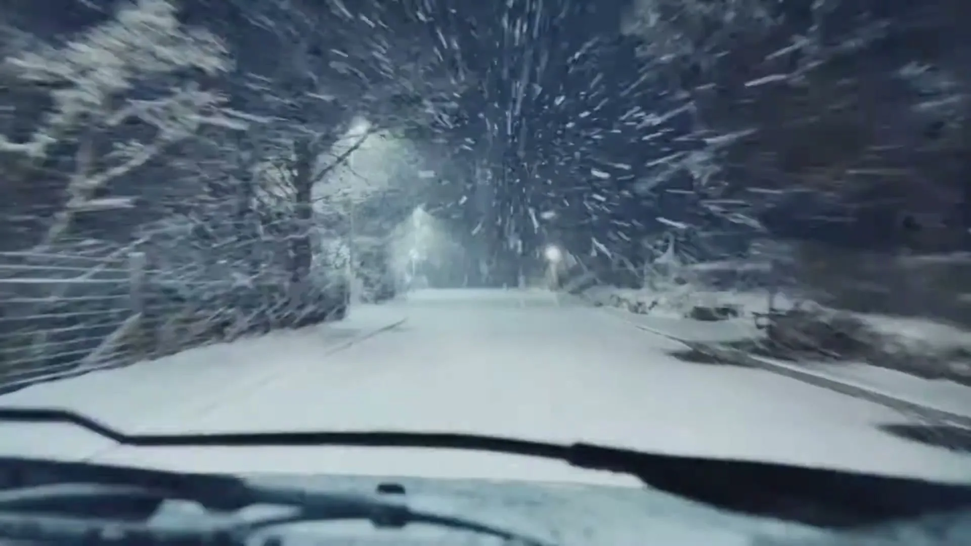 A car drives down a snow-covered road at night, with streetlights illuminating the falling snow and trees lining the path.