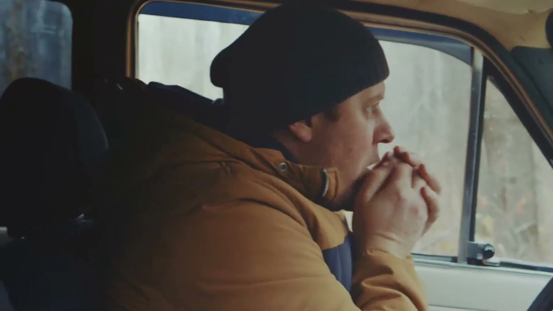A man in a beanie and a brown jacket blows on his hands to warm them while sitting in a car.