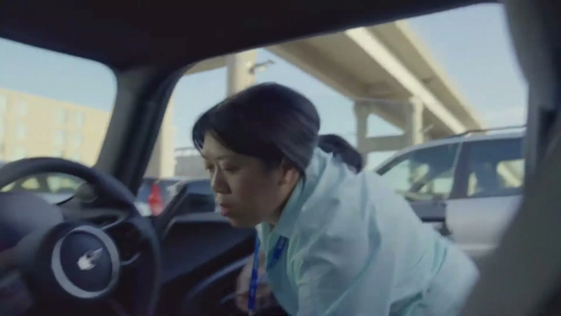 A woman in a light blue shirt leans into a car, looking for something on the floor, in a parking lot under an overpass.