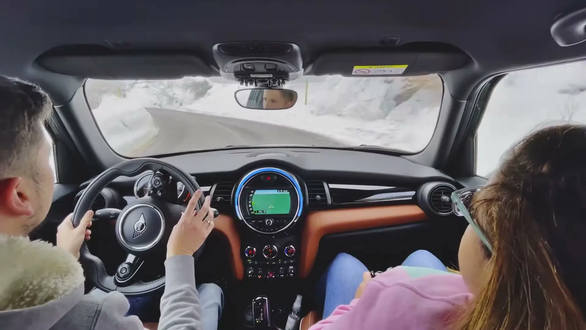 A couple drives a Mini Cooper on a snowy mountain road, seen from the back seat, with the driver's hands on the wheel and the passenger looking out the window.