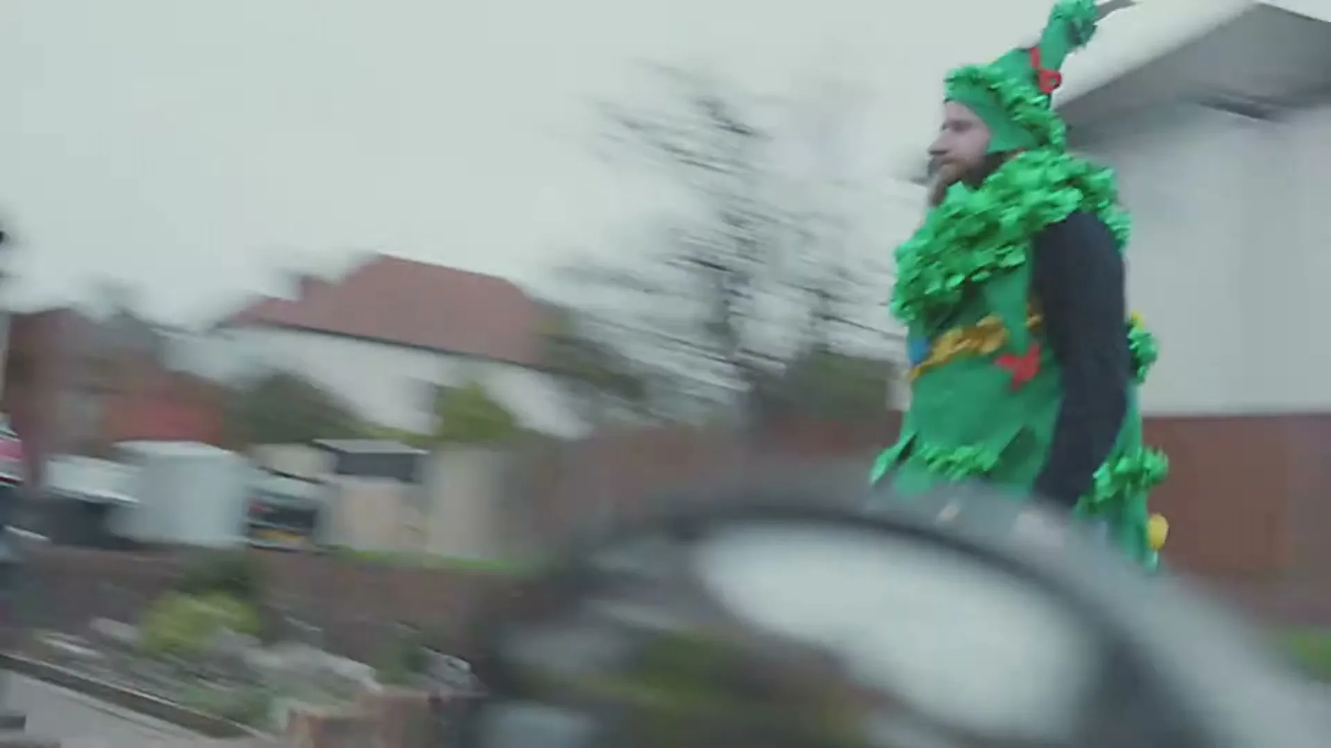 A man in a Christmas tree costume walks past a car on a suburban street during the day.