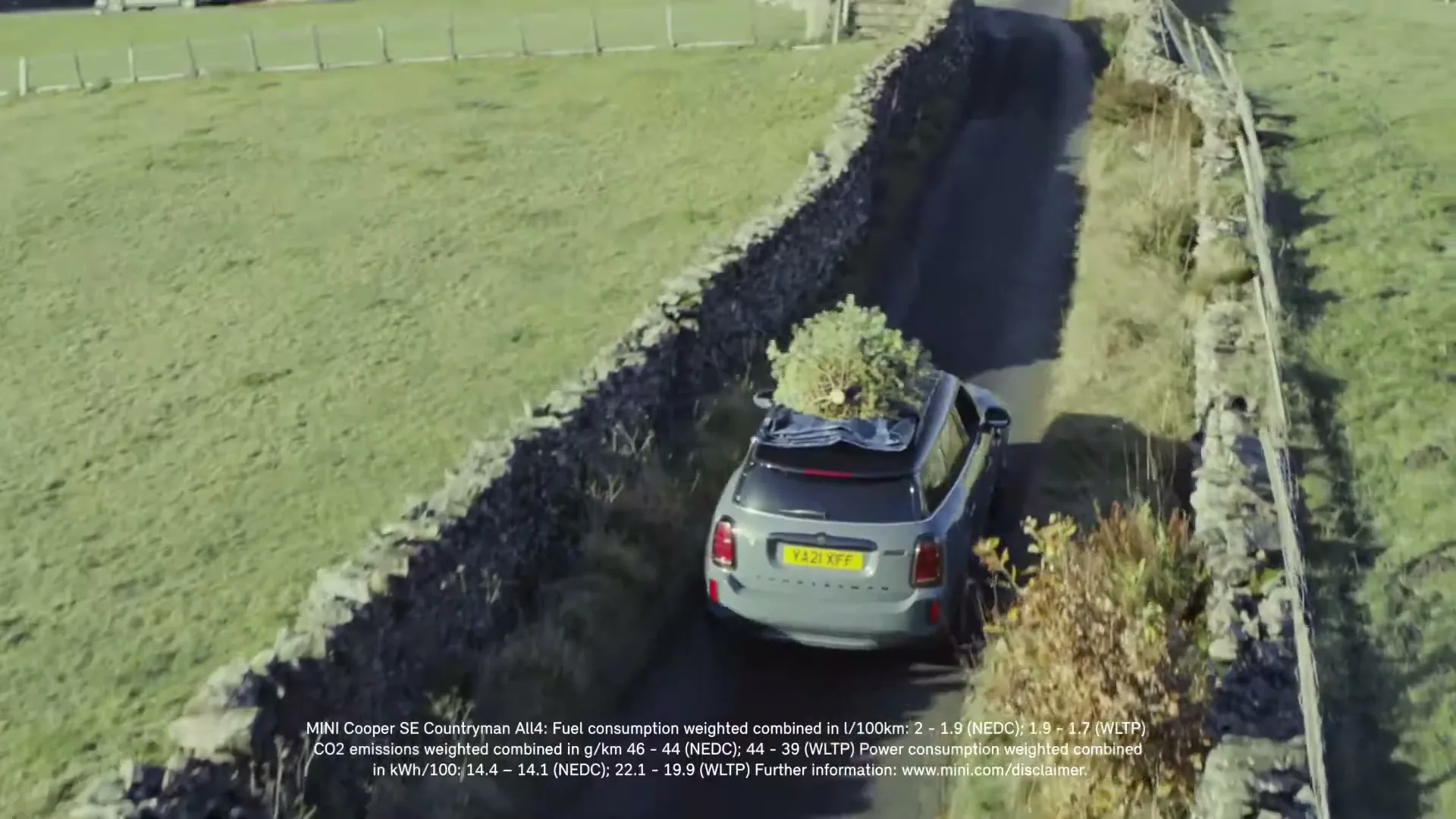 A grey Mini Cooper SE Countryman All4 drives down a narrow country road with a Christmas tree on its roof, surrounded by stone walls and green fields.