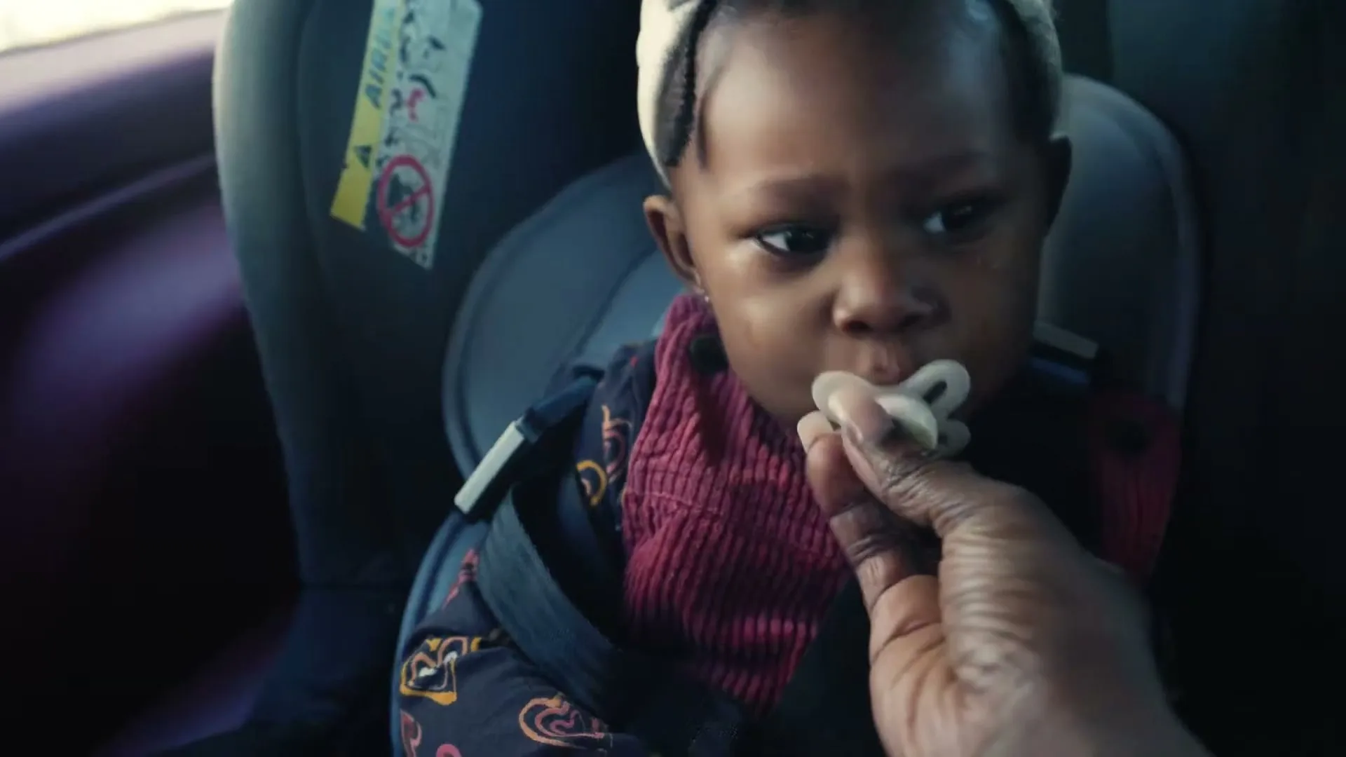 A baby in a car seat cries as an adult hand offers a pacifier, inside a car during the day.