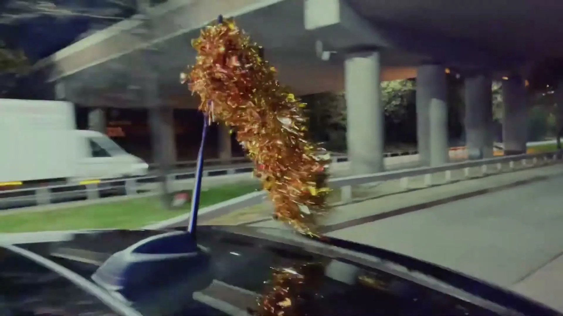A close-up shot of a car's antenna decorated with gold tinsel, driving at night under an overpass.