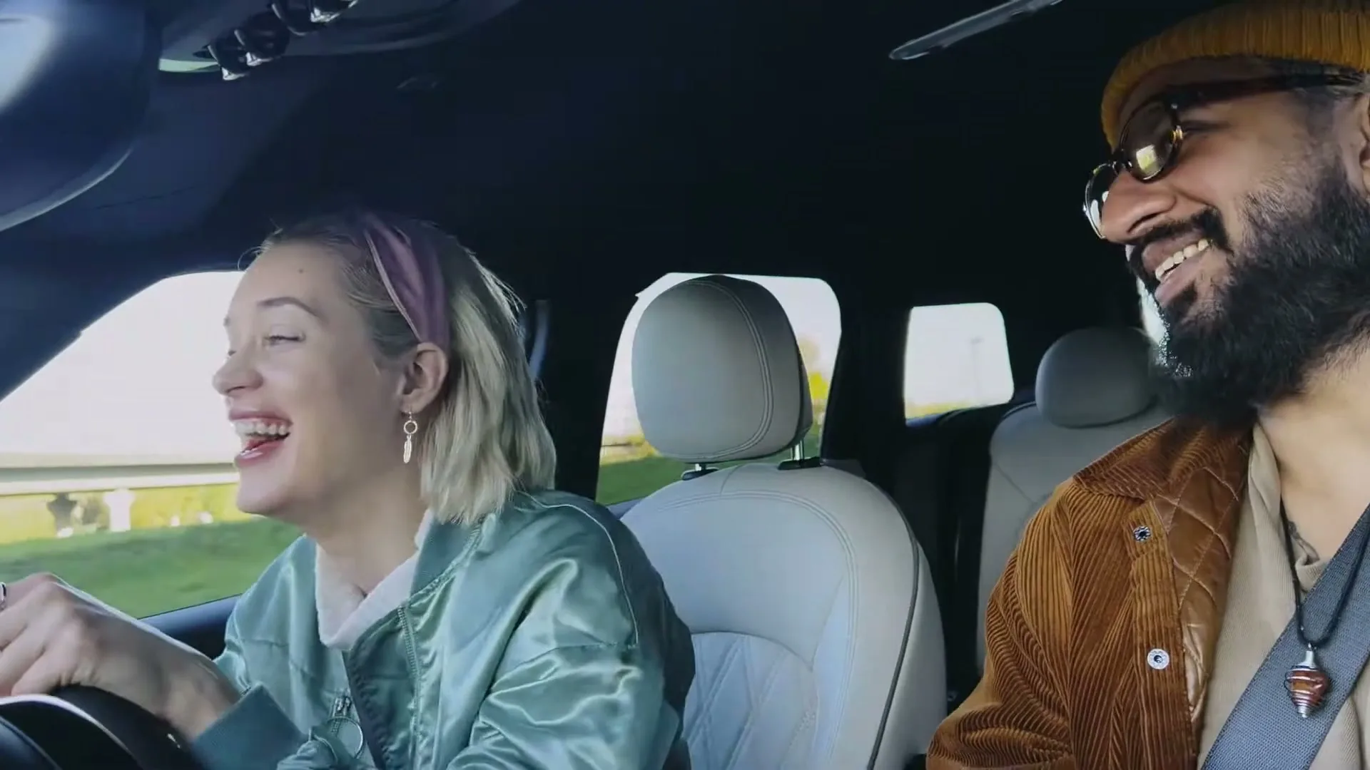 A young blonde woman in a green jacket and a man with a beard and beanie laugh while driving a car on a sunny day.