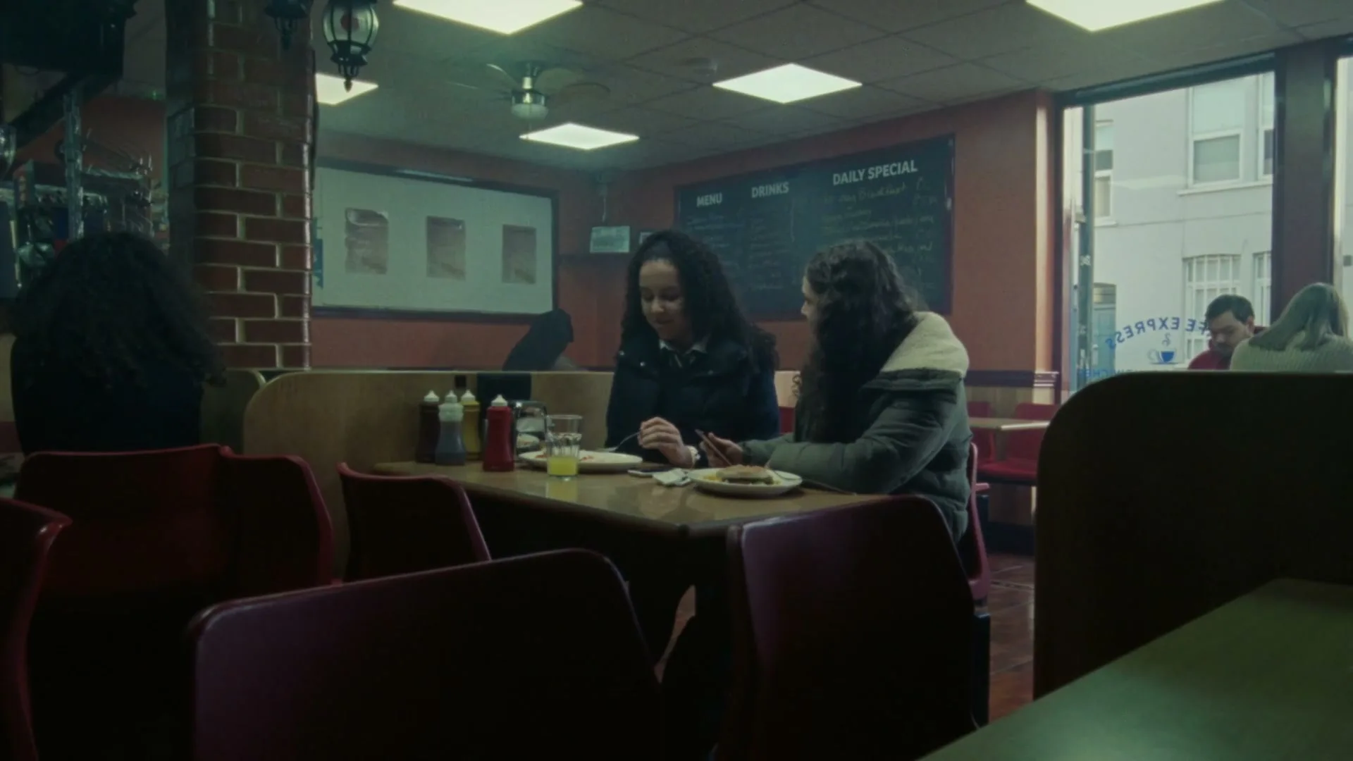 Two young women with dark curly hair sit at a booth in a diner, talking and looking at a phone.