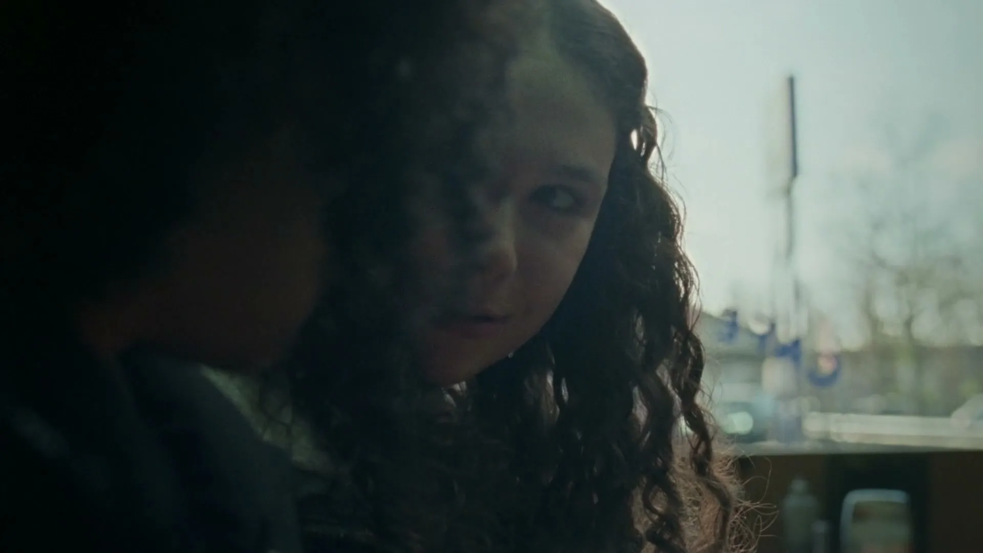 A young woman with curly hair looks over her shoulder at someone off-screen, with a blurred exterior visible through a window behind her.