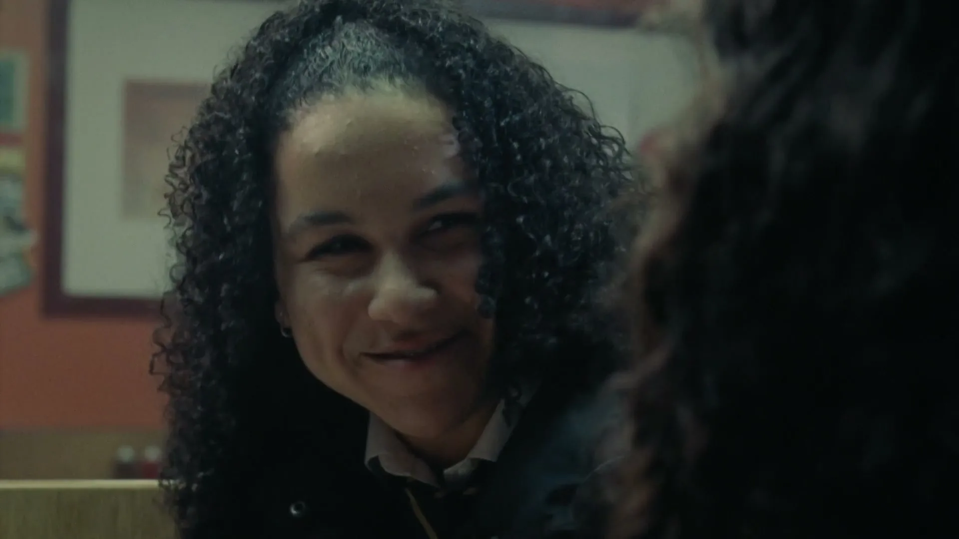 A young woman with curly hair and braces smiles mischievously at someone off-screen in a dimly lit interior setting.
