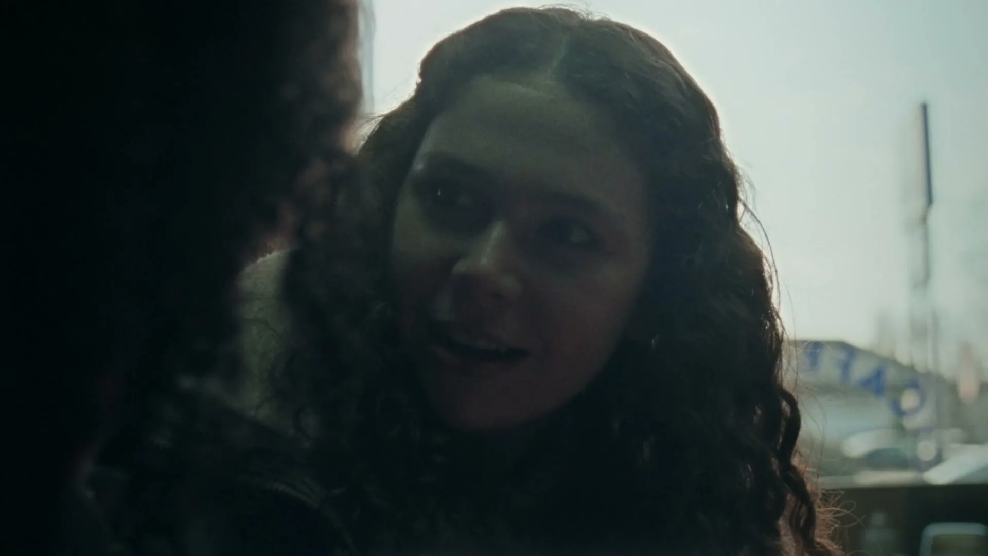 A young woman with long, curly dark hair talks to someone off-screen, her expression shifting from surprise to a slight smile, in a dimly lit interior.
