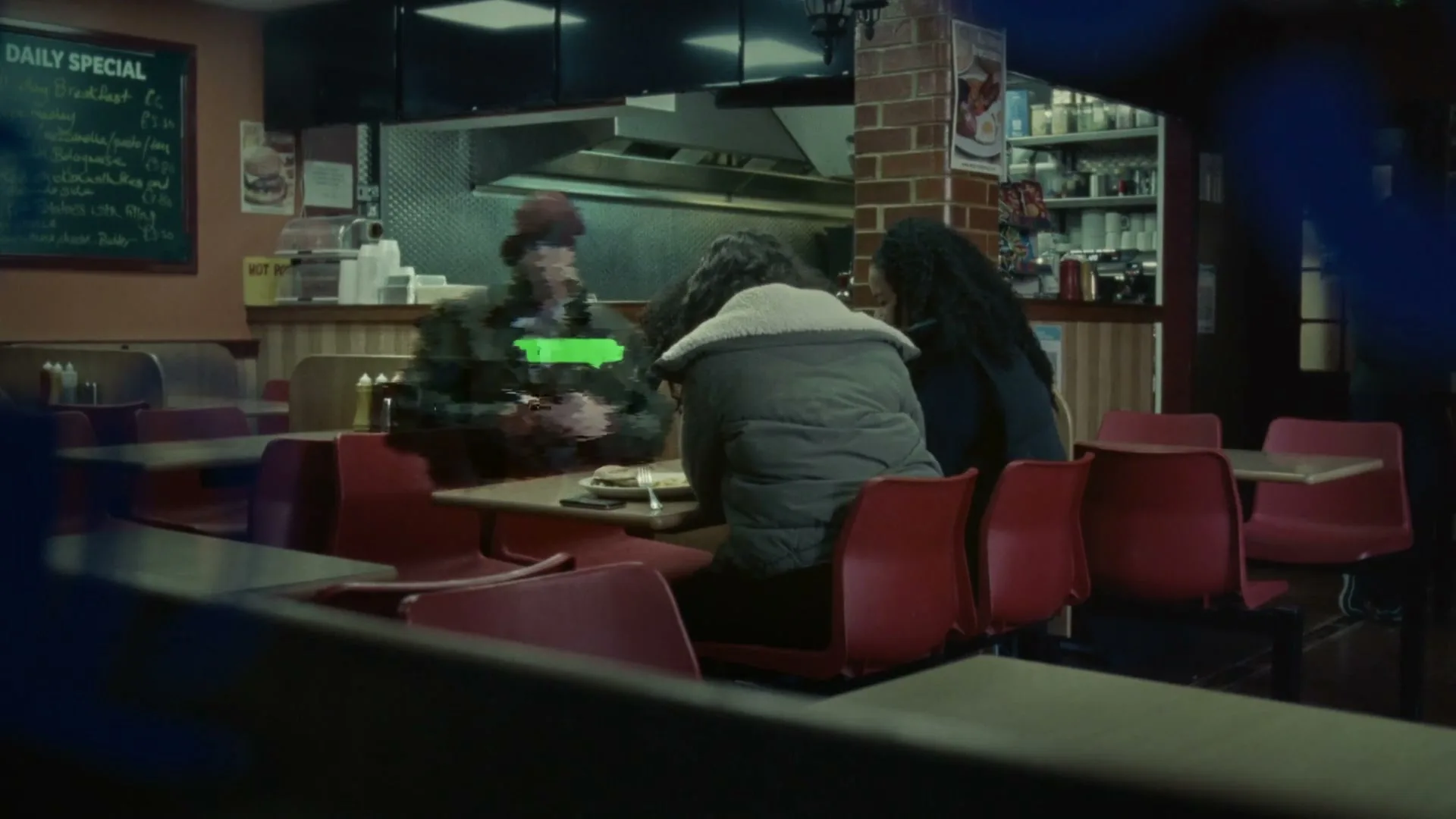 Two women and a man sit at a table in a diner, with the man appearing distorted and glitching.