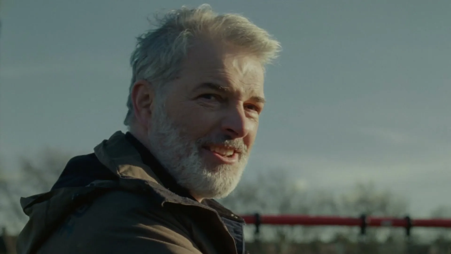 A middle-aged man with a grey beard and a brown jacket turns his head to look at the camera and smiles outdoors.