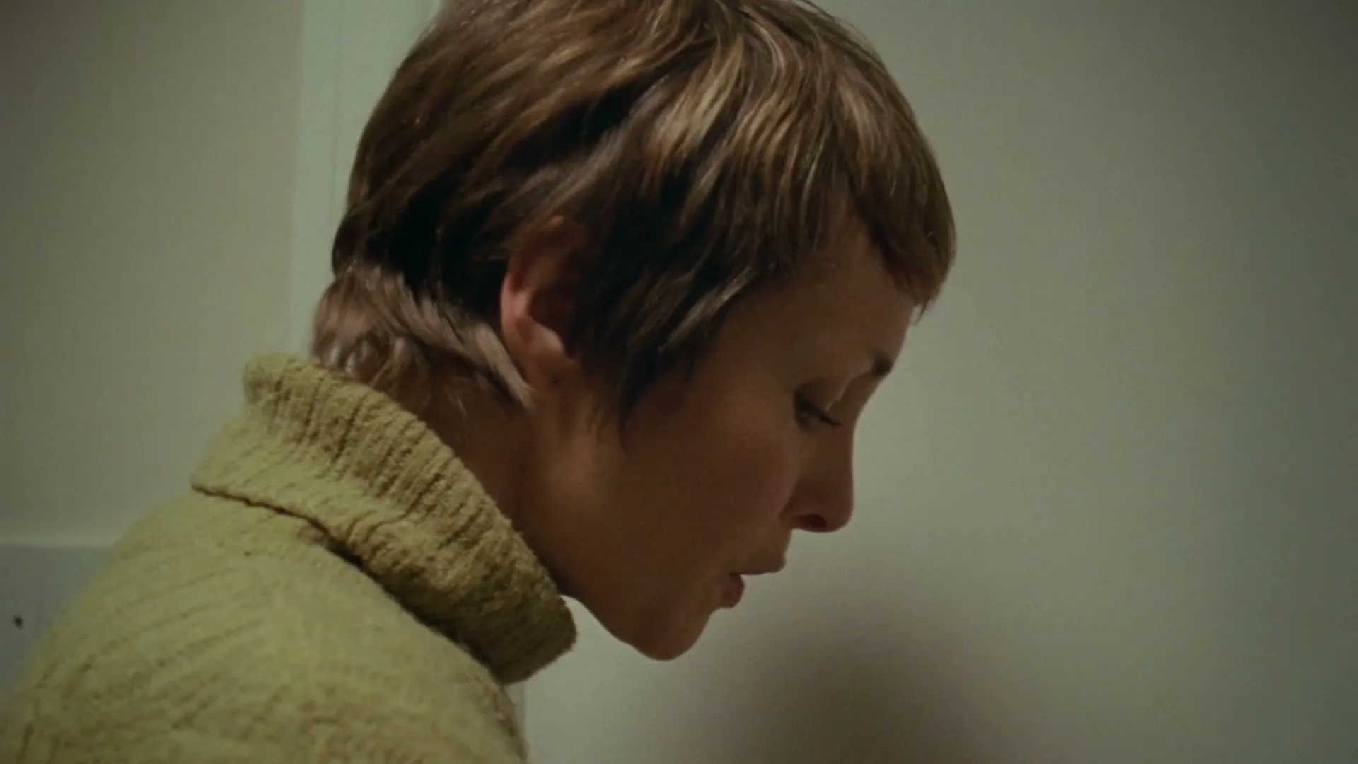 A woman with short brown hair and a yellow turtleneck sweater looks down, her mouth slightly open, in a dimly lit room.