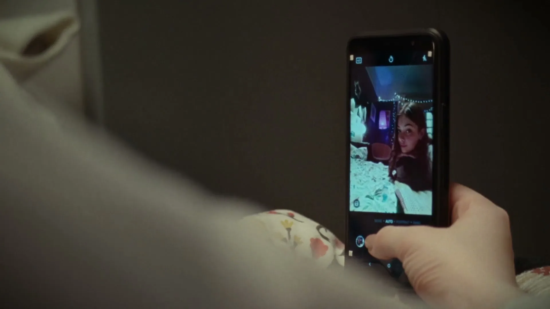 A person's hand holds a smartphone, displaying a selfie of a young woman in a dimly lit room with string lights, as the person takes a photo.