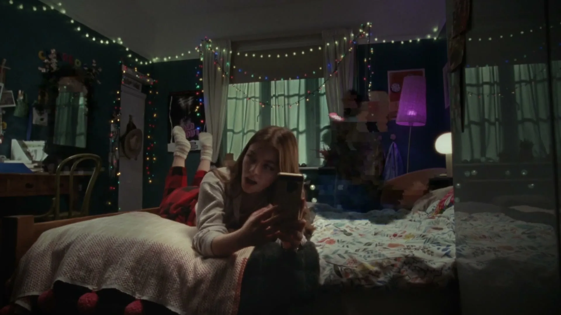 A young woman with long brown hair lies on her bed, looking at her phone, in a dimly lit bedroom decorated with string lights.