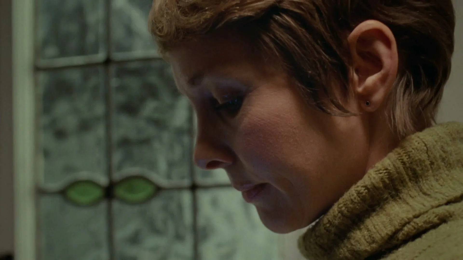 A woman with short brown hair and a green sweater looks down with a concerned expression, next to a stained glass window.