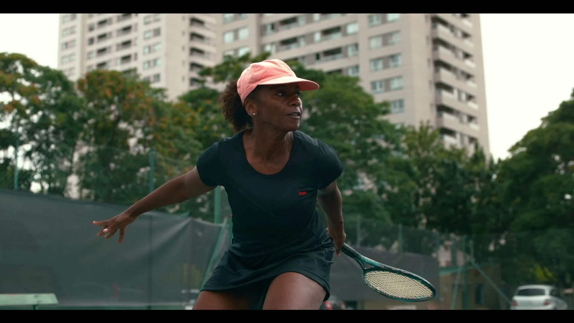 A female tennis player in a black outfit and pink visor is poised to hit a tennis ball on an outdoor court, with apartment buildings in the background.