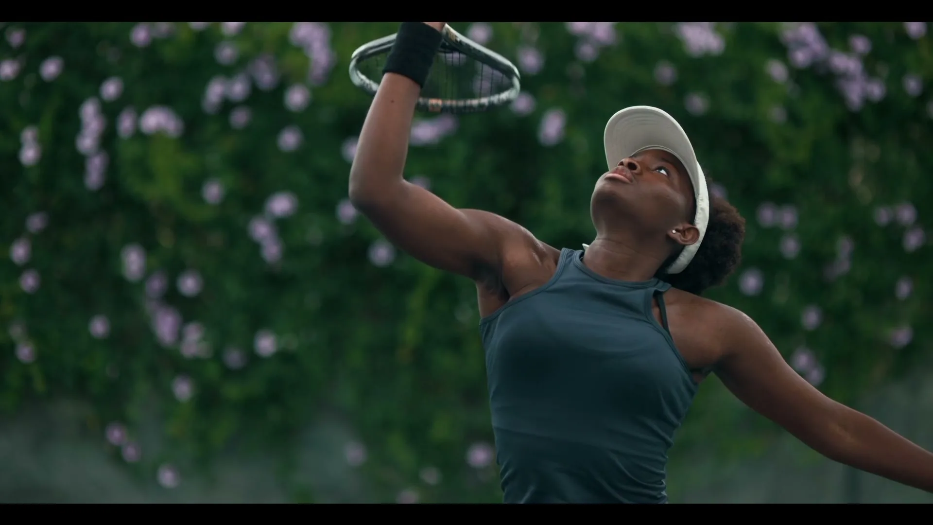 A young Black female tennis player in a dark tank top and white visor serves on a tennis court with green foliage in the background.