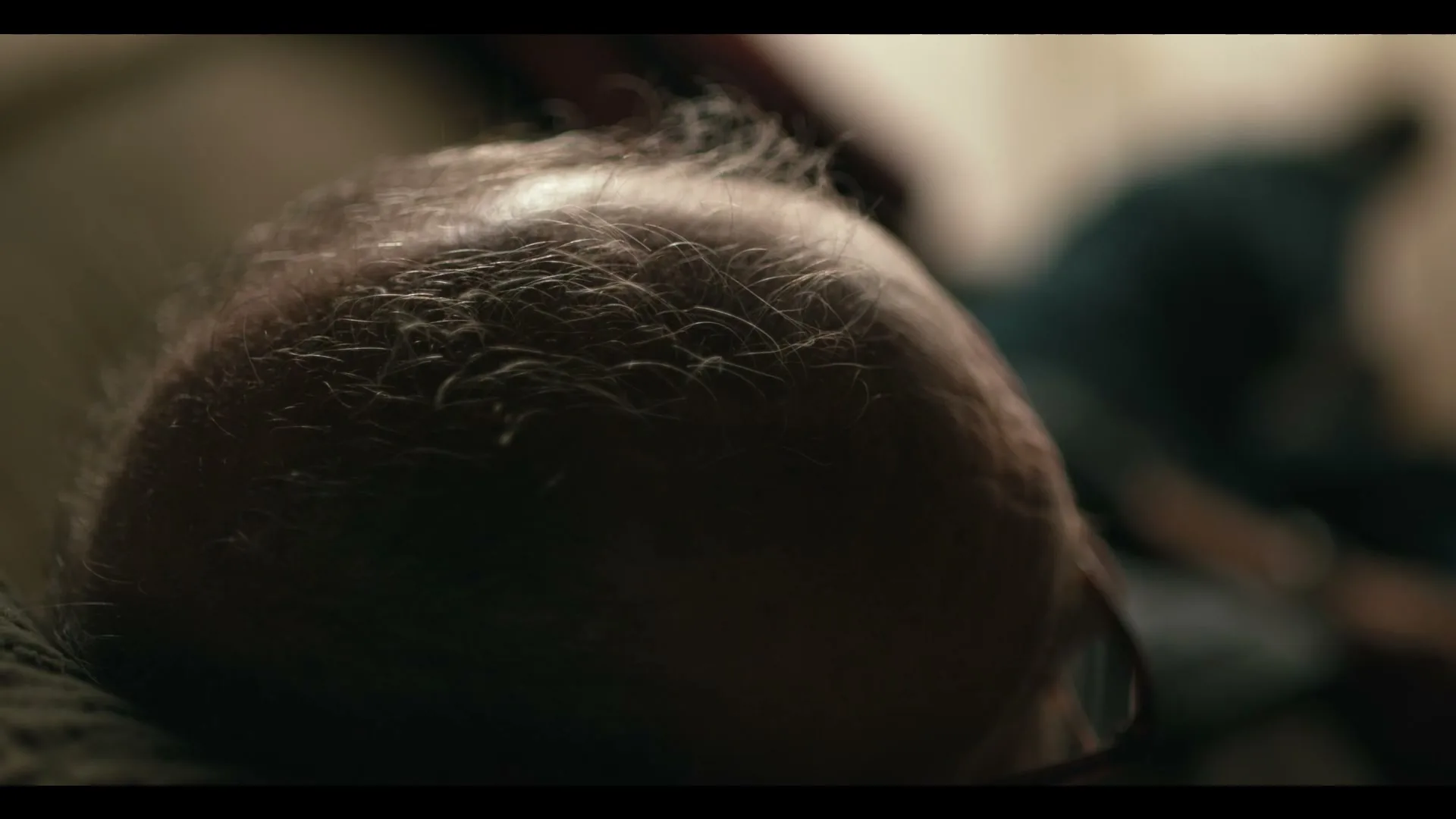 A man with thinning hair lies on a pillow, seen from an overhead perspective, in a dimly lit interior setting.