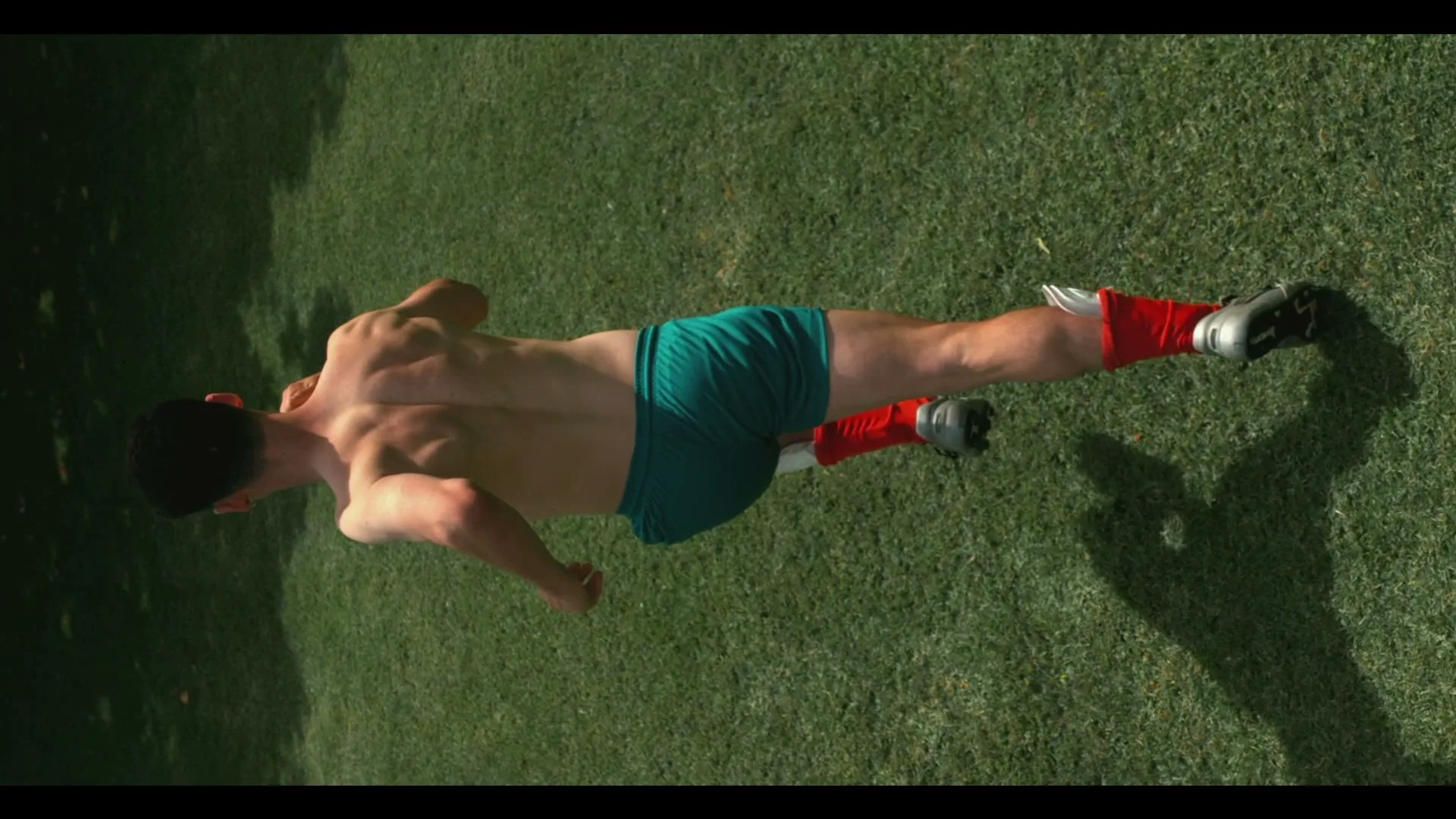 A shirtless young man with a prosthetic leg runs across a grassy field, viewed from directly above, with sunlight and shadows.
