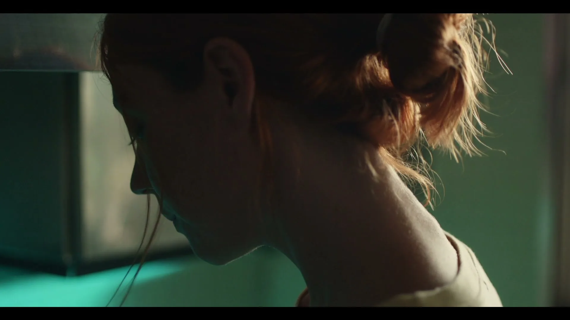 A young woman with red hair in a bun looks down, illuminated by a green light in a laboratory setting.