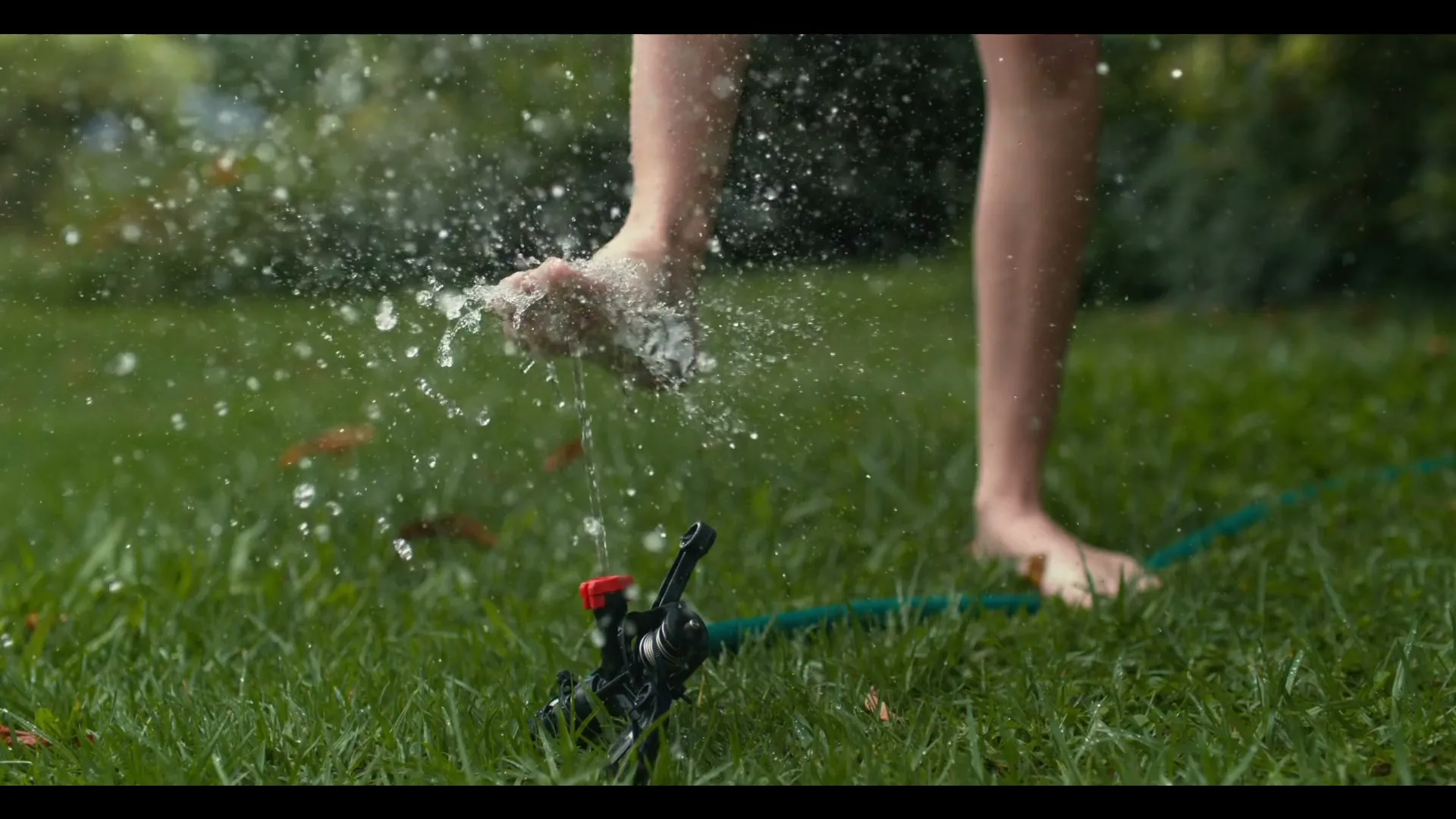 A child's bare feet play in the water from a sprinkler on a green lawn, splashing water droplets in the air.