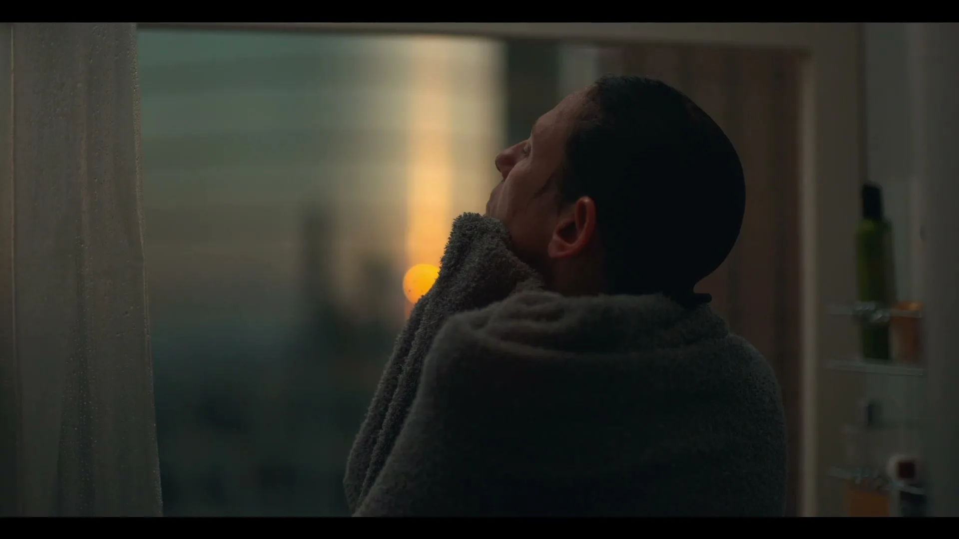A man with short dark hair, wrapped in a grey towel, looks up and to the right, with a blurry sunset visible through a window in the background.