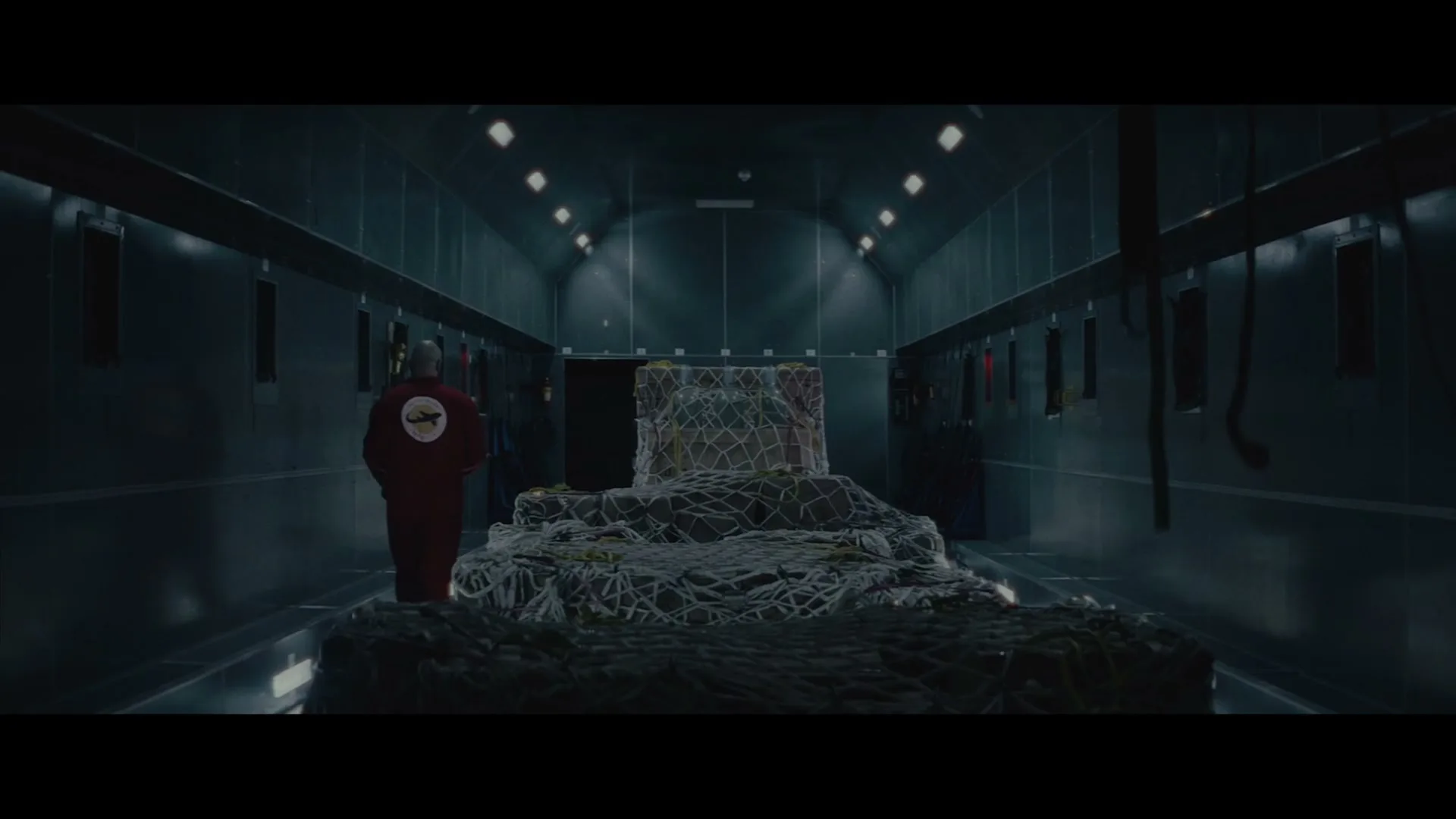 A man in a red jumpsuit with a logo on his back stands in a dark, metallic cargo hold, looking at a large, netted package.