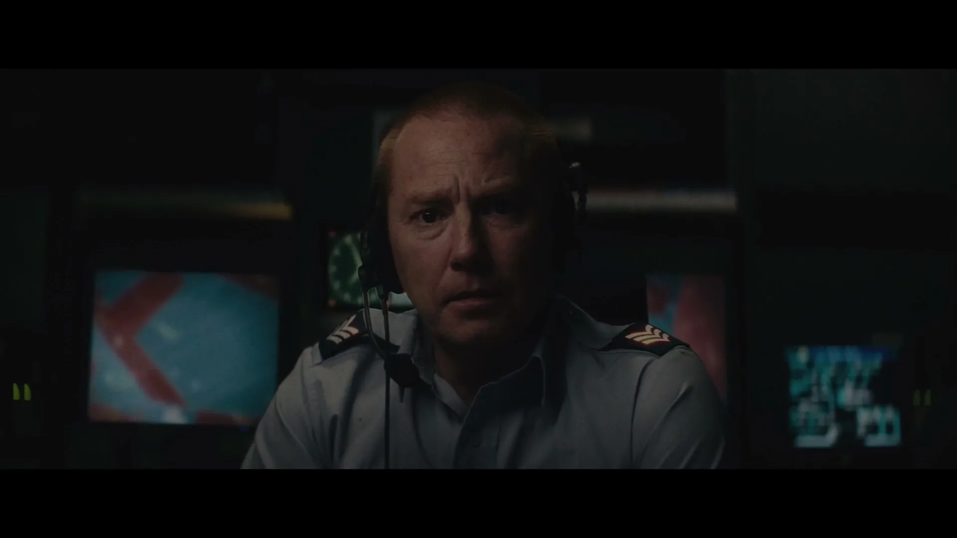 A man in a military uniform and headset looks directly at the camera in a dark control room with multiple screens.