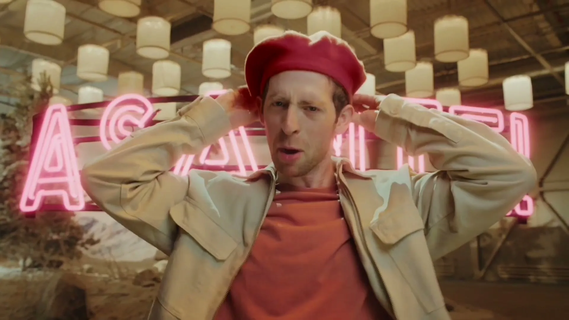 A man in a red beret and beige jacket smiles and gestures with his hands in front of a pink neon sign that reads "ASANTE!" in a studio set.