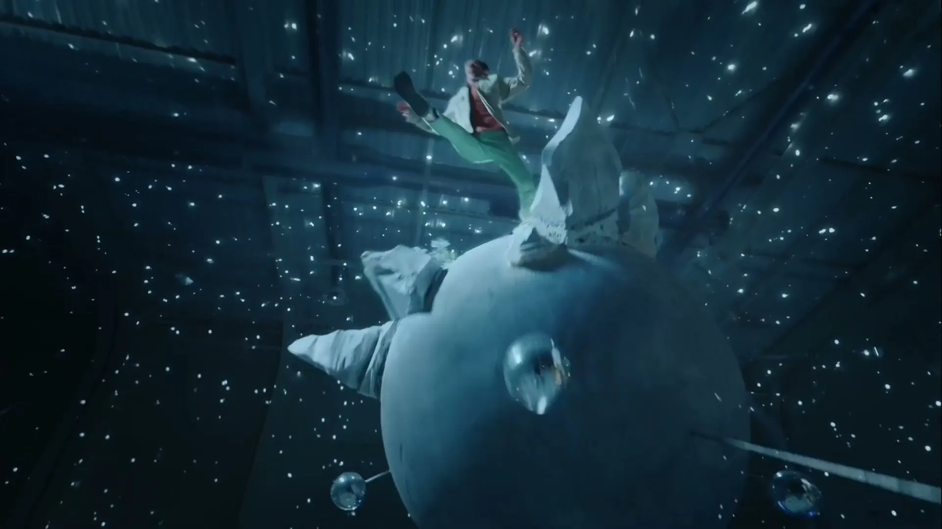 A young man in a suit stands on a large, round, white object, then jumps off as small, reflective spheres float around him in a dark, starry space.